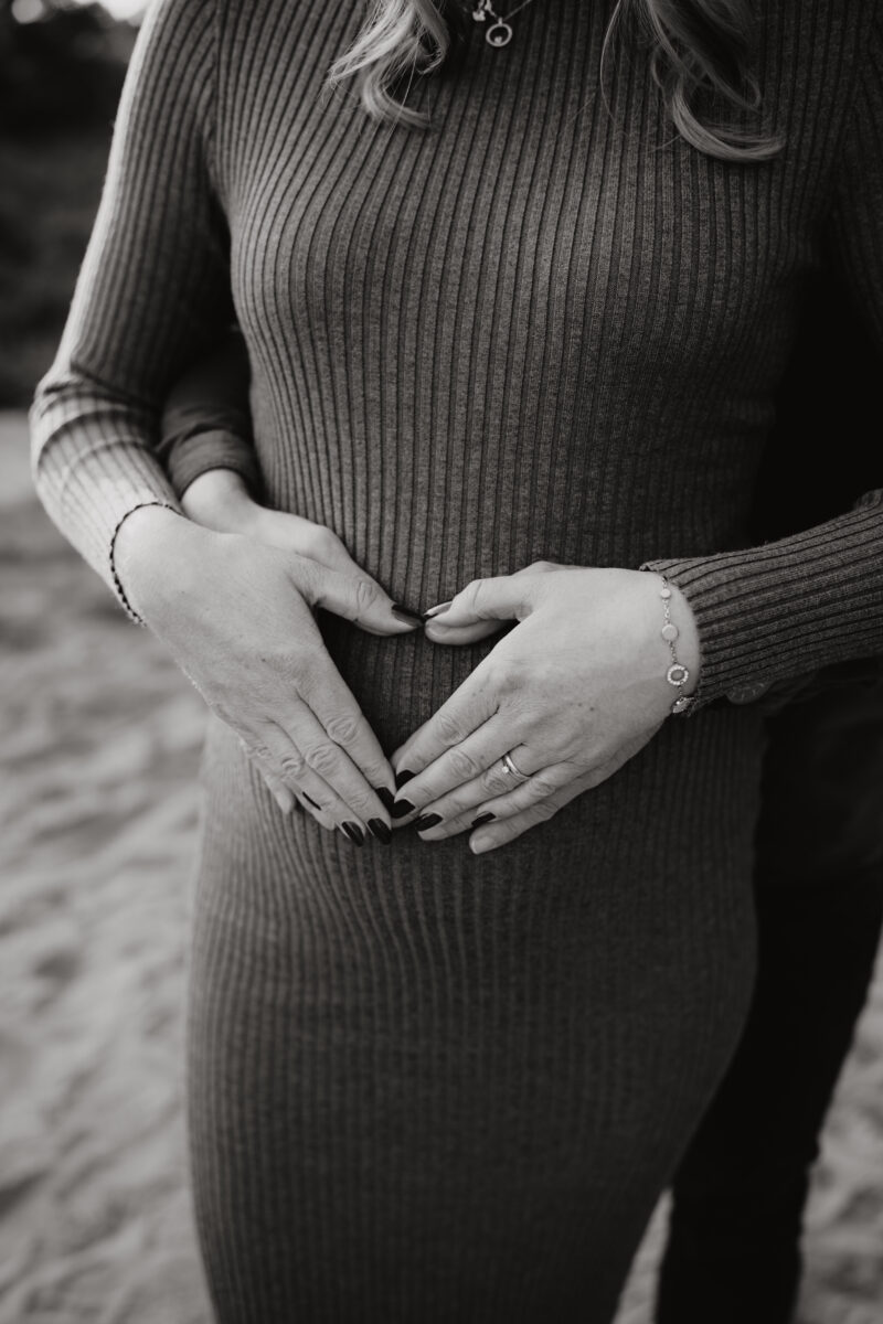 Babybauch Fotografie Portfolio Nahaufnahme vom Babybauch und den Händen der Eltern die ein Herz formen Schwarz-Weiß-Fotografie