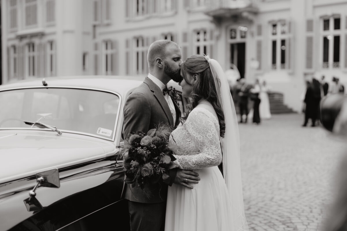 Hochzeitslocation, Braut und Bräutigam stehen gemeinsam vor einem Auto, der Bräutigam küsst die Braut auf die Stirn, Bild in Schwarz Weiß