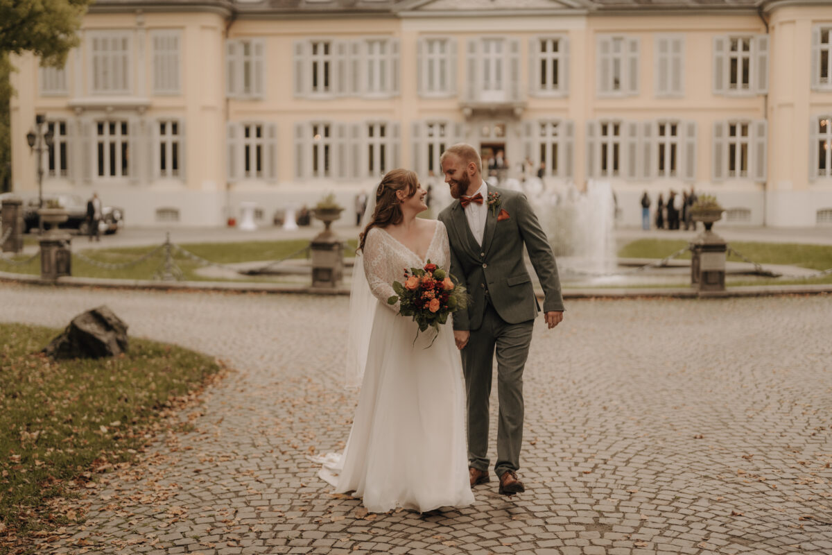 Hochzeitsportfolio, Brautpaar vor einem Schloss und einem Brunnen, gemeinsam am gehen und schauen sich lächelnd an