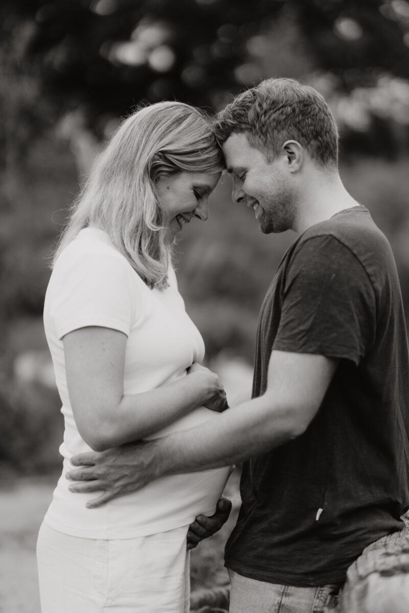 Babybauch Fotografie Portfolio glückliches junges Paar schaut auf den Bauch der werdenden Mutter Outdoor Schwarz-Weiß-Fotografie