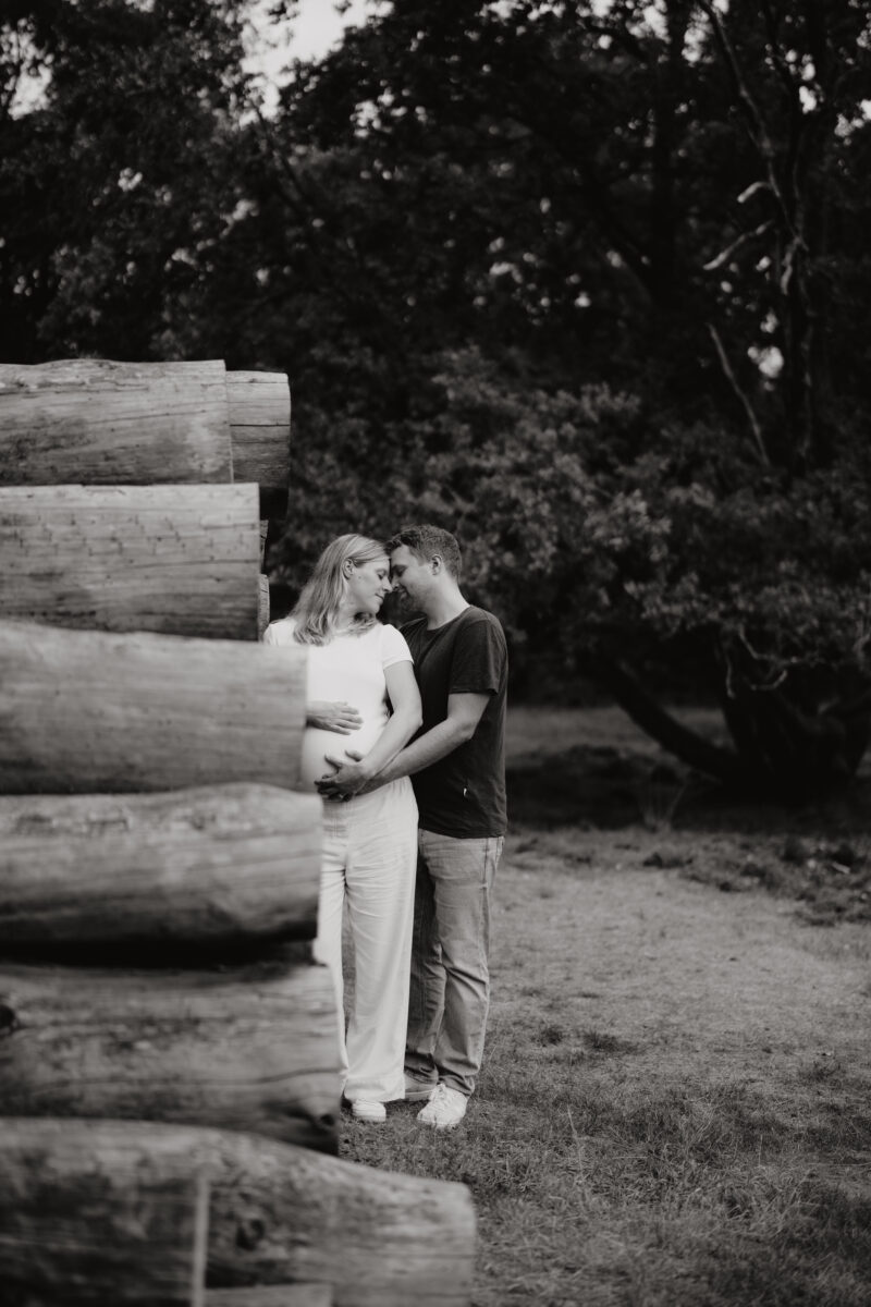 Babybauch Fotografie Portfolio glückliches junges Paar hinter aufgestapeltem Holz Outdoor Schwarz-Weiß-Fotografie