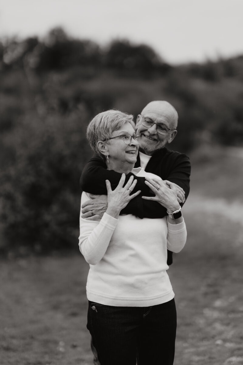 Er umarmt sie von hinten und lächelt, fotografiert in Schwarz-Weiß