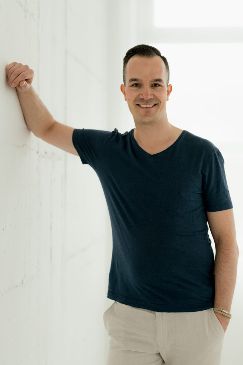 Portraitfoto eines Mannes, an einer Wand lehnend, lächelnd in die Kamera