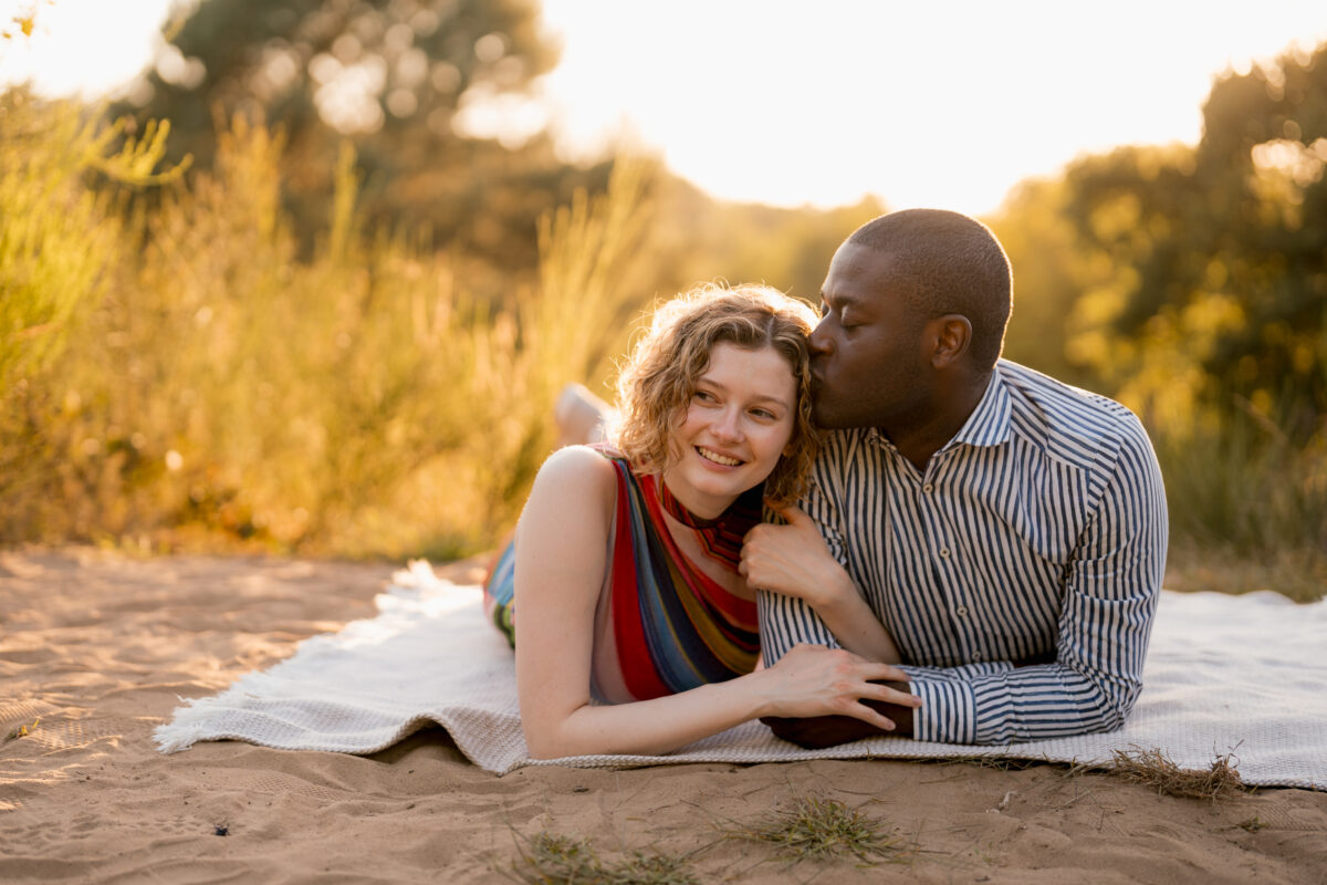 Paar liegt in der Abendsonne auf einer Decke und sie bekommt einen Stirnkuss