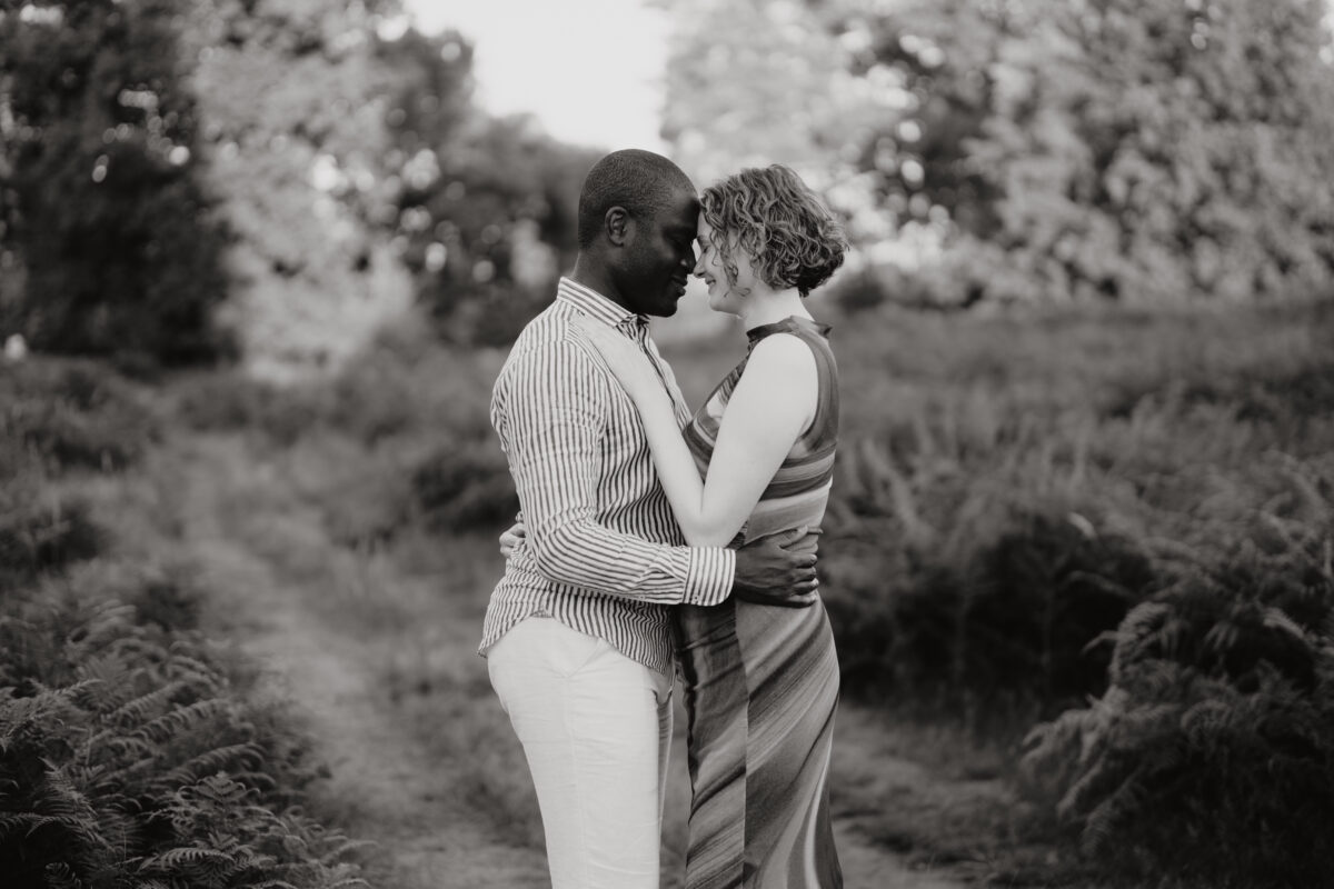 Paar steht Stirn an Stirn im Wald und lächelt, fotografiert in Schwarz-Weiß