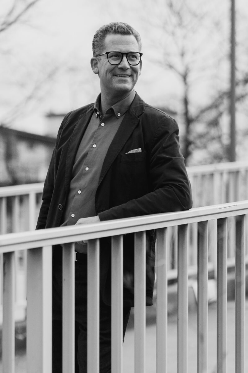 Schwarz-Weiß Portraitfoto eines Mannes, wie er sich an ein Geländer anlehnt