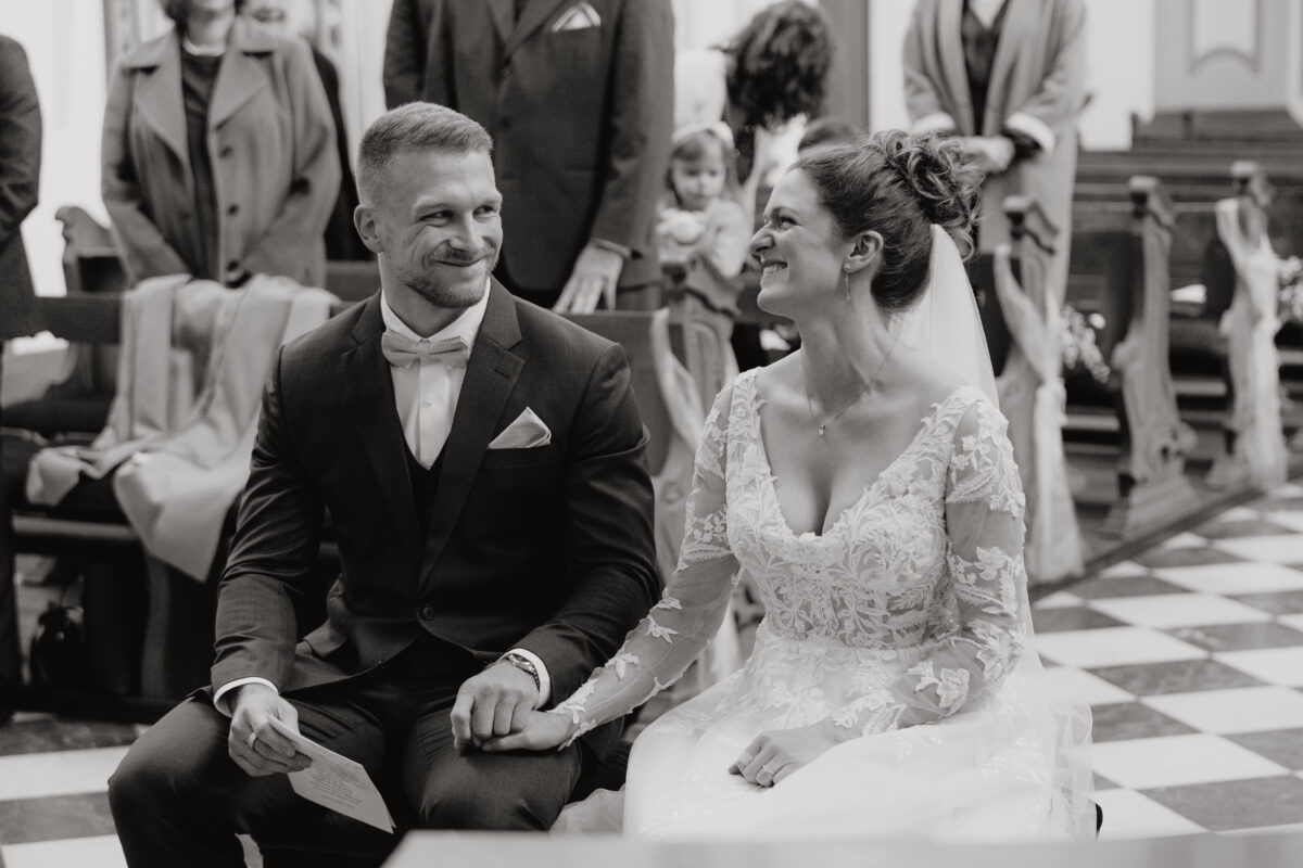 Hochzeitsportfolio, Hochzeitspaar sitzen vorne, lachen sich herzlich an, Bräutigam hält die Hand der Braut fest