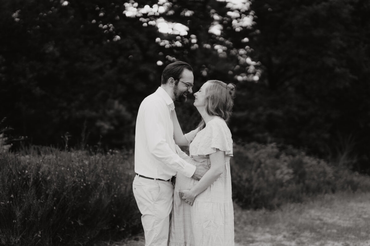 Babybauch Fotografie Portfolio glückliches junges Paar steht vor einer Wiese und schaut sich an Outdoor Schwarz-Weiß-Fotografie