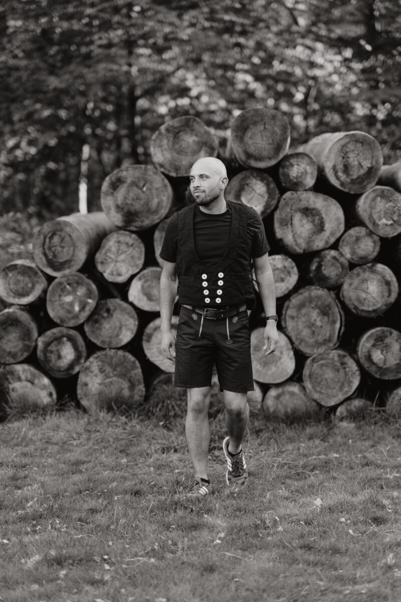 Schwarz-Weiß Portraitfoto eines Mannes vor gestapelten Baumstämmen, in Tischlerkluft