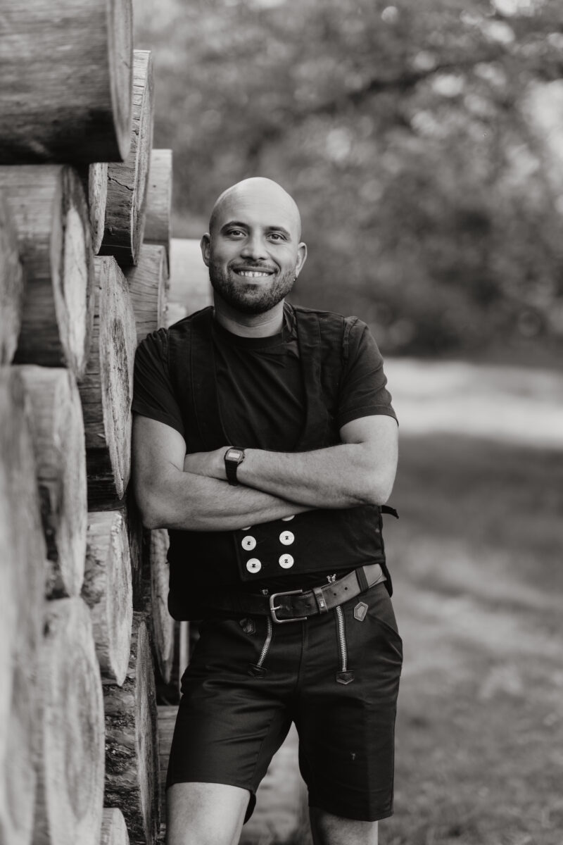 Schwarz-Weiß Portraitfoto einer Mannes in Tischlerkluft, wie er sich an aufgestapelte Baumstämme lehnt