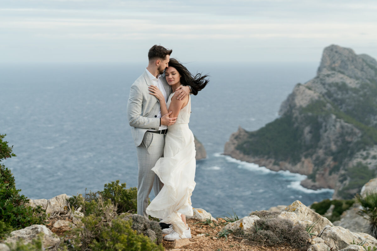 Hochzeitsportfolio, Braut lehnt sich an Bräutigam und beide kuscheln im Hintegrund felsige Klippen und das Meer