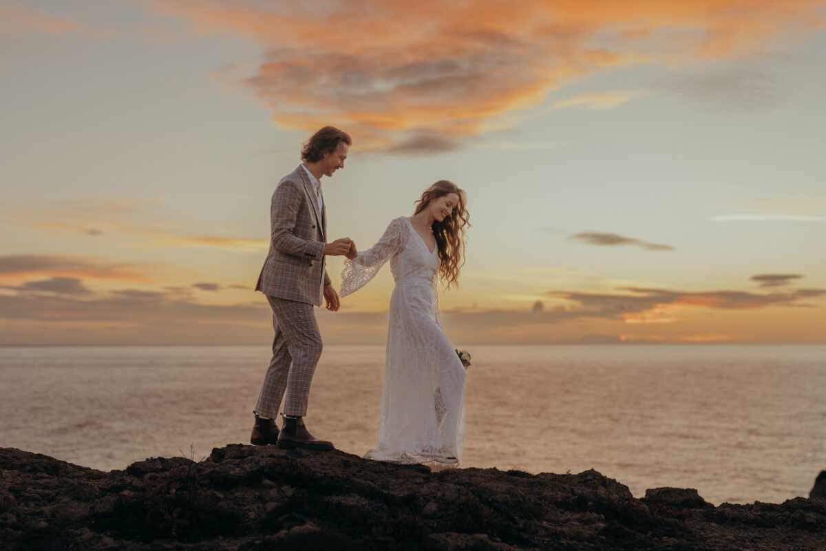 Hochzeitsportfolio, Braut und Bräutigam laufen Hand in Hand die steinige Klippe entlang im Hintergrund die untergehende Sonne und das Meer