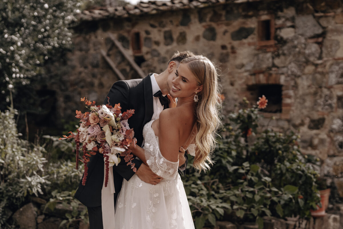 Destination Wedding Portfolio Hochzeitspaar umarmt sich mit Brautstrauß vor verschwommenem Hintergrund