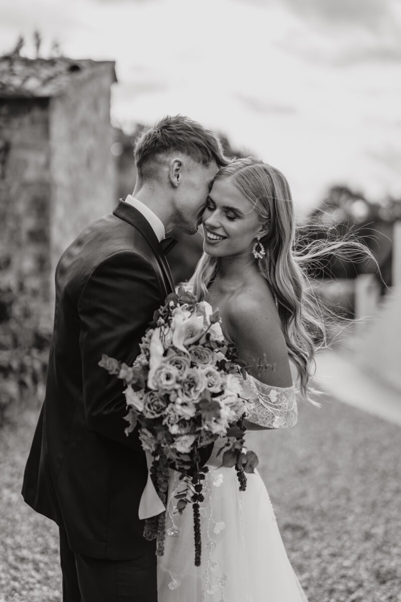 Destination Wedding Portfolio Hochzeitspaar umarmt sich mit Brautstrauß vor verschwommenem Hintergrund, schwarz-weiß