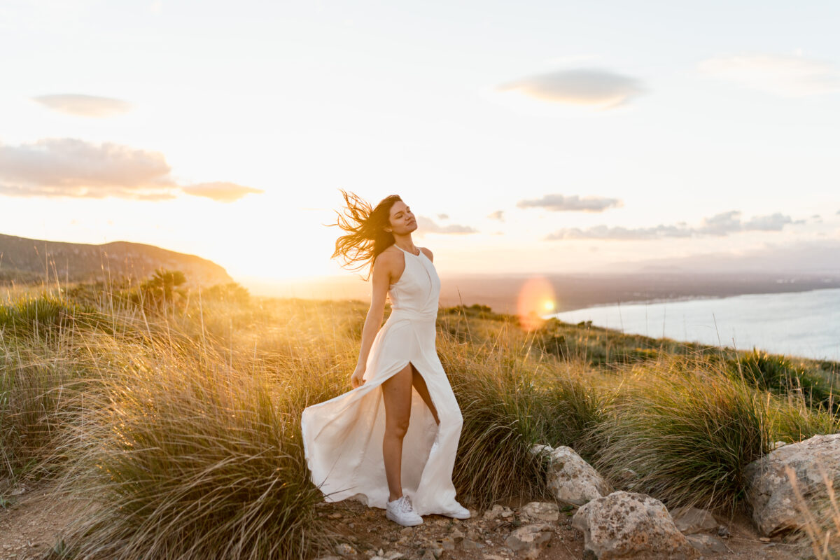 Destination Wedding Portfolio Braut in wehendem weißen Kleid auf einer Wiese vor untergehender Sonne