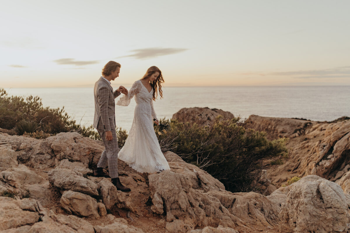 Destination Wedding Portfolio Hochzeitspaar hält sich an den Händen und steigt über Felsen am Meer
