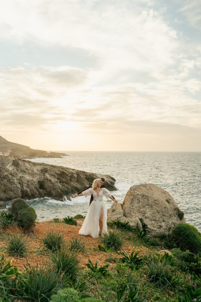 After Wedding, Totale, Braut und Bräutigam stehen vor der Felsenlandschaft und dem Meer. Der Bräutigam hält die Hände seiner Braut von hinten, beide haben die Arme ausgestreckt.