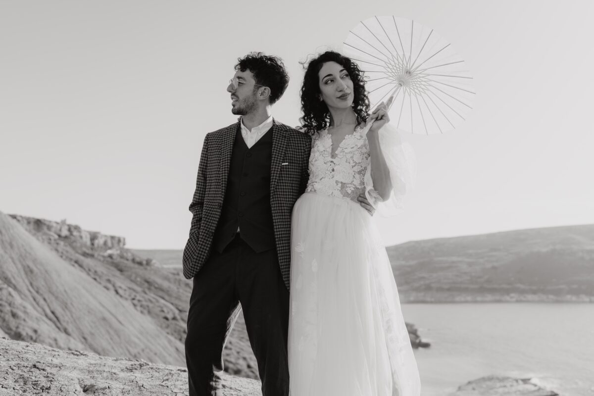 After Wedding Portfolio, Braut und Bräutigam halten sich im Arm. Die Braut hat in der rechten Hand einen Sonnenschirm, den sie über sich hält. Braut und Bräutigam schauen in andere Richtungen in die Ferne.