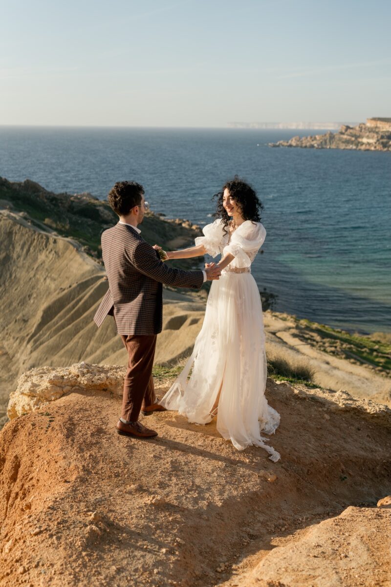 After Wedding Portfolio, Braut und Bräutigam halten sich an den ausgrstreckten Händen beide schauen sich lächelnd an. Beide stehen auf der Höhe eines Berges, im Hintergrund sieht man das Meer