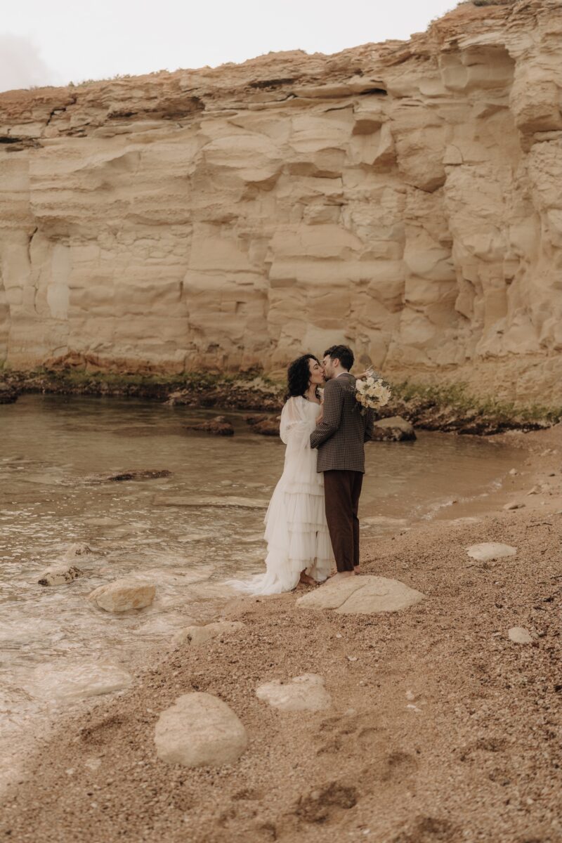 After Wedding Portfolio, Braut und Bräutigam küssen sich am Wasser, Umgeben von einer Felsenlandschaft. Beide halten sich aneinander fest.