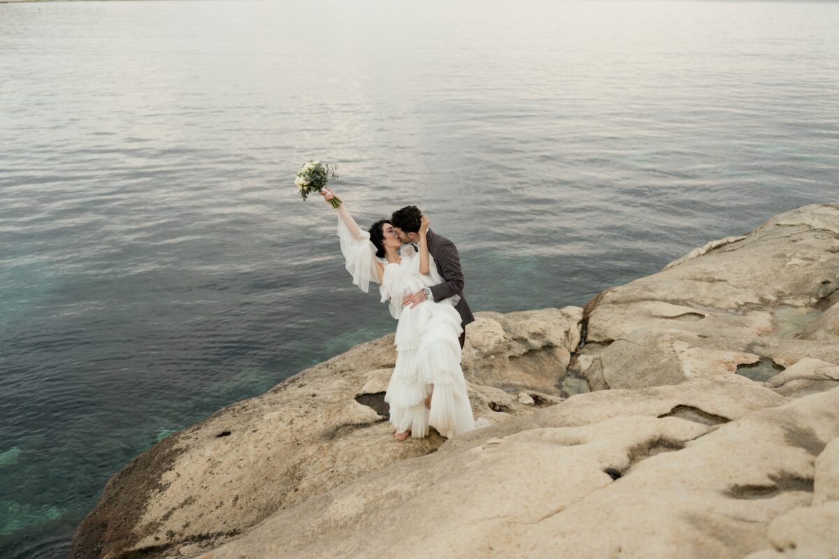 After Wedding Portfolio, Braut und Bräutigam stehen auf einem Felsen im Hintergrund ist das Meer. Der Bräutigam lehnt die Braut nach hinten. Sie hält sein Gesicht mit der Hand und hält die linke Hand mit dem Blumenstrauß nach oben