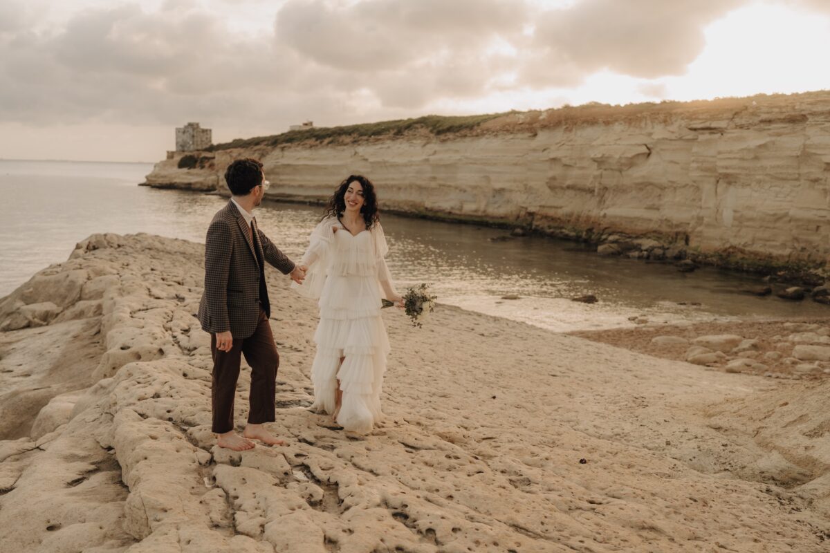 After Wedding, Braut und Bräutigam schauen sich beide an und halten sich an der Hand. Sie hält ihren Brautstrauß in der Hand. Beide laufen am Meer entlang.