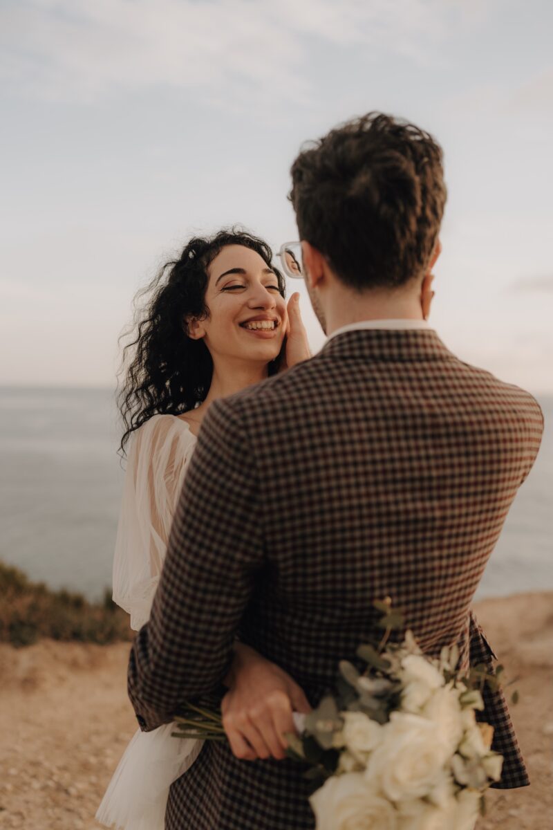 After Wedding Portfolio, Braut und Bräutigam stehen sich gegenüber. Der Bräutigam mit dem Rücken zu der Kamera. Die Braut lächelt ihn an. Erhät ihr Gesicht mit der Hand. Im Hintergrund ist das Meer zu sehen.