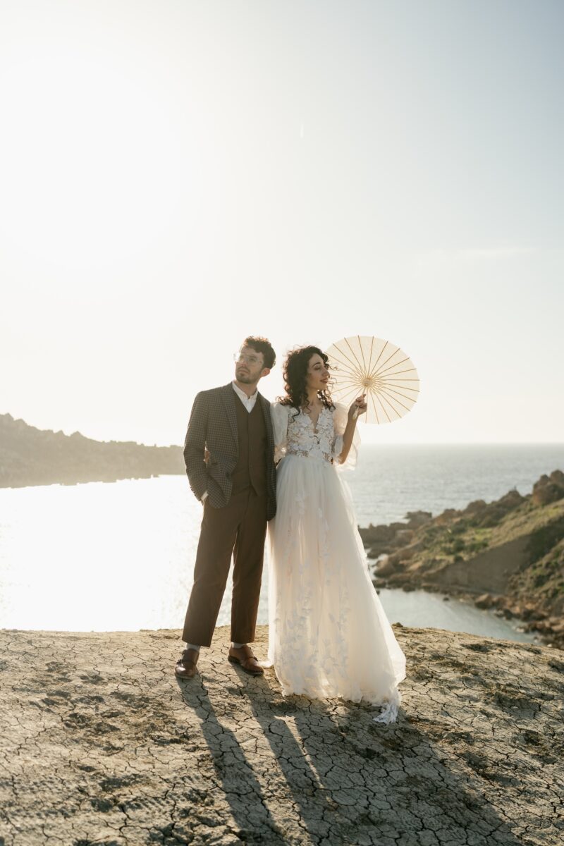 After Wedding Portfolio, Braut und Bräutigam blicken die die Ferne. Die Braut nach rechts, der Bräutigam nach links. Beide stehen nebeneinander. Er hat die Hände in den Hosentaschen. Sie hält einen Sonnenschirm.