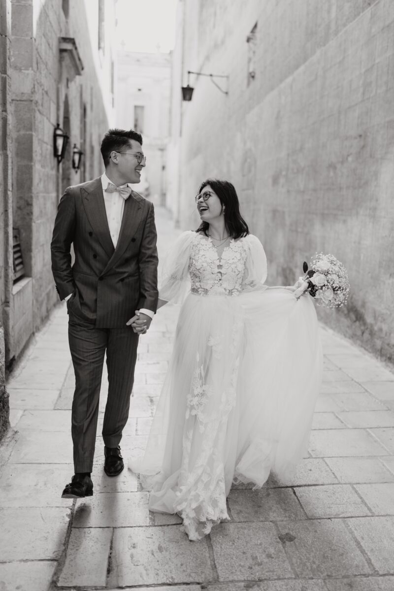 After Wedding, Braut und Bräutigam stehen in einer Gasse. Sie halten sich an der Hand und schauen sich gegenseitig an. In der anderen Hand hält sie den Brautstrauß und er hat die Hand in seiner Hosentasche.