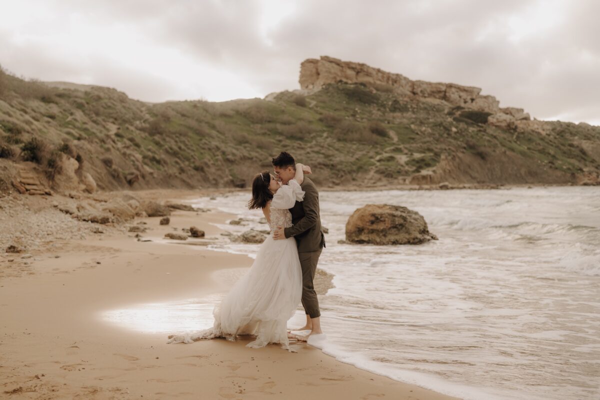 After Wedding Portfolio, Braut und Bräutigam stehen vor einer Felsenlandschaft, direkt am Meer. Das Wasser umspühlt das Brautkleid. Braut hat die Arme um den Nacken des Bräutigams geschlungen. Beide schauen sich tief in die Augen.