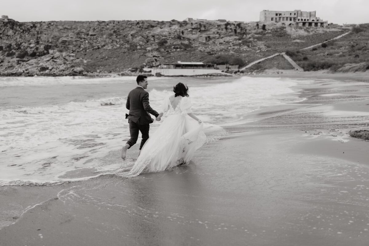 After Wedding Portfolio, Braut und Bräutigam laufen zusammen durch die Gischt des Meeres, sie halten sich dabei an der Hand. Man sieht beide von hinten.