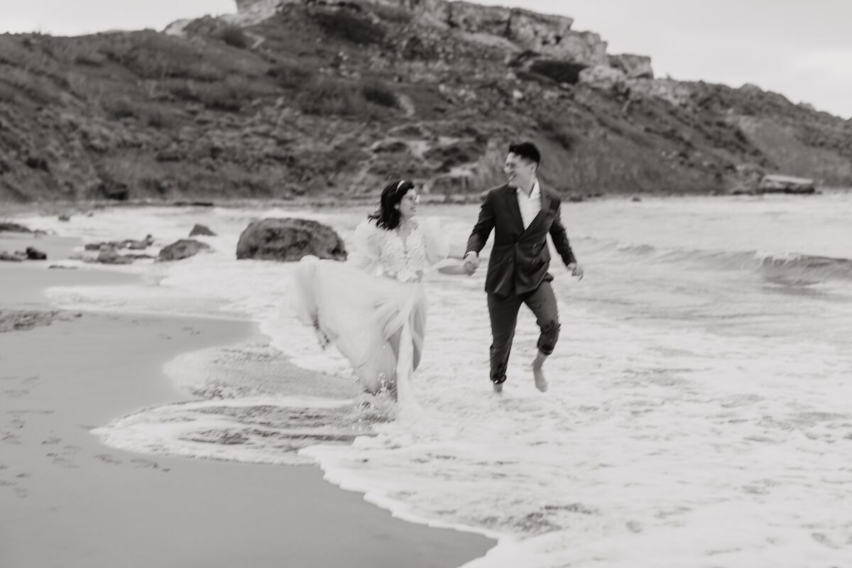 After Wedding Portfolio, Braut und Bräutigam sind beide am Strand und laufen in der Gischt. Sie schauen sich beide lächelnd an. Im Hintergrund ist ein großer Felsen