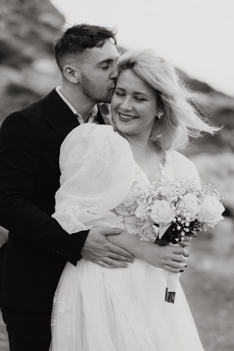 After Wedding, der Bräutigam umarmt seine Braut von der Seite und küsst sie seitlich auf die Stirn. Sie lächelt leicht nach links und hält ihren Brautstrauß