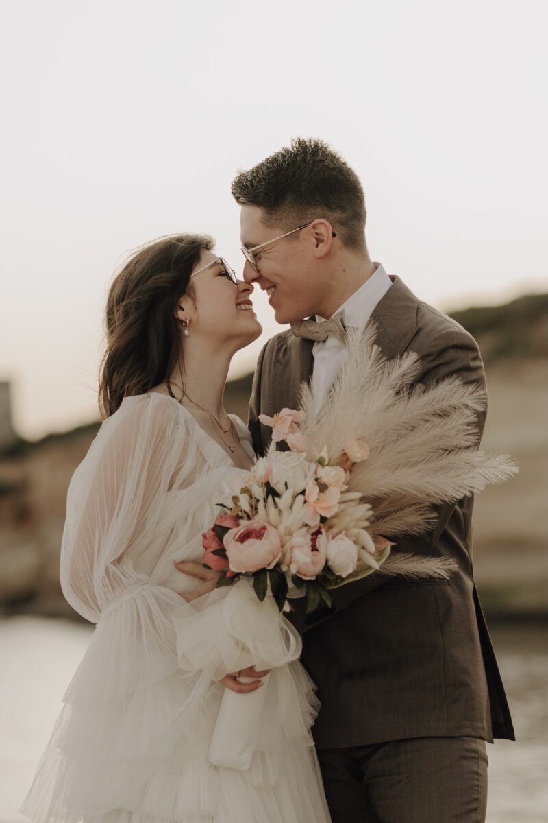 After Wedding, Braut und Bräutigam lachen sich an, ihre Nasenspitzen berühren sich. Sie hält ihren Brautstrauß