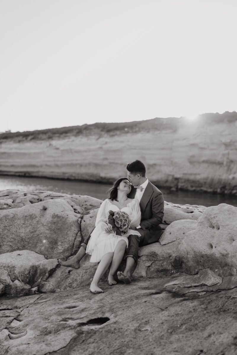 After Wedding, Braut und Bräutigam sitzen auf einem Felsen. Sie lehnt sich an ihn. Beide schauen sich lächelnd an