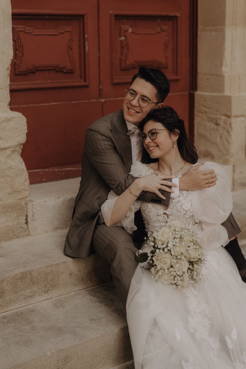 After Wedding, Braut und Bräutigam sitzen auf einer Treppe. Die Braut liegt in den Armen des Bräutigams und er umarmt sie von hinten. Beide lächeln und schauen in die Ferne.
