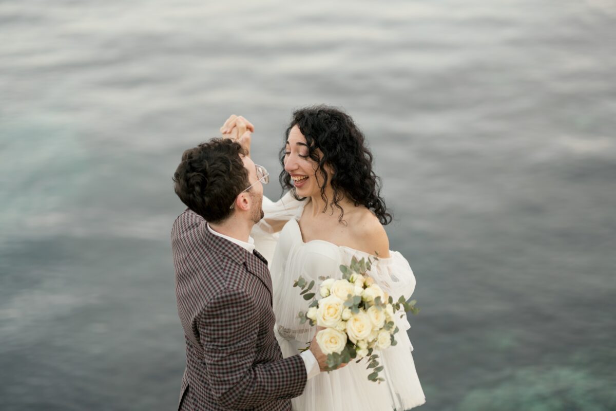 After Wedding Portfolio, Braut und Bräutigam lächeln sich an ihre Hände sind ineinander verschränkt. Im Hintergrund sieht man das Wasser, des Meeres