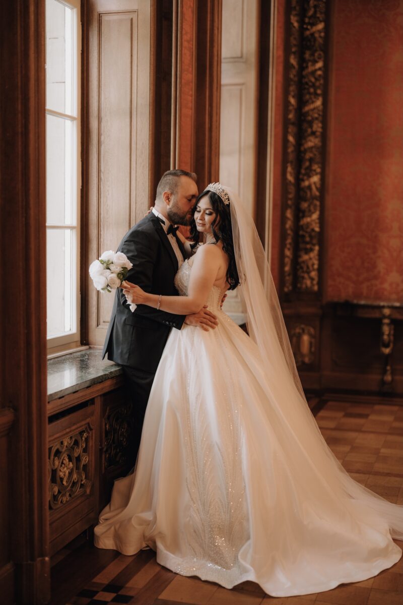 After Wedding Portfolio, der Bräutigam hält die Taille der Braut und küsst ihre Wange. Braut hält den Strauß in der linken Hand und lächelt zur Seite.