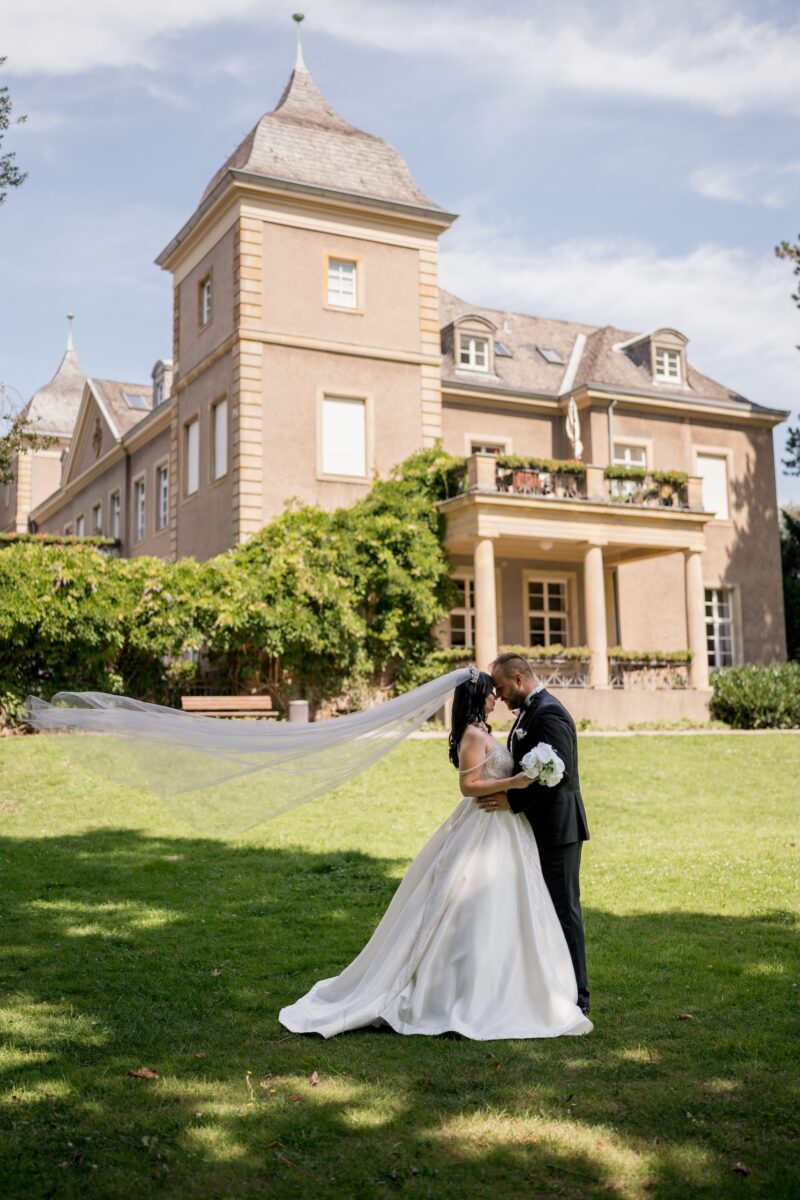 After Wedding Portfolio, Braut und Bräutigam stehen vor einem Haus. Ihre Köpfe liegen aneinander und der Schleier, der Braut weht nach hinten. Beide halten sich aneinander fest.