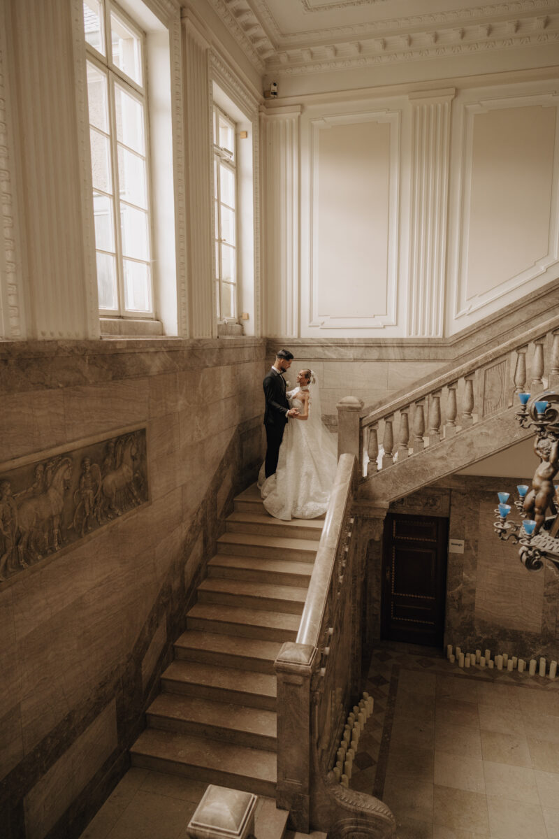 Hochzeitsportfolio, Braut und Bräutigam stehen auf einer Mamortreppe, innerhalb eines Gebäudes. Er hält sie an der Taille fest. Beide schauen sich lächelnd an.