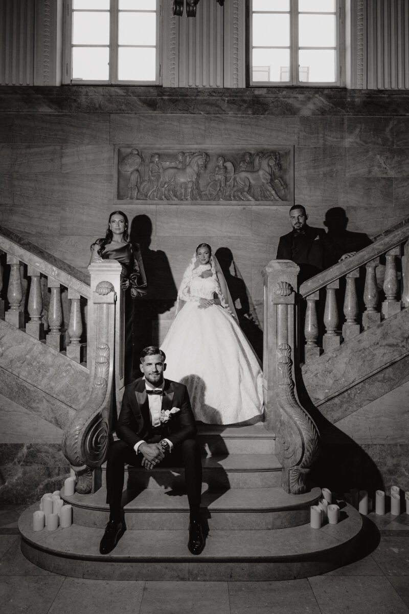 Hochzeitsportfolio, Gruppenfoto angeblitzt auf einer Mamor treppe, Bräutigam sitzt auf der untersten Treppe und hat die Arme auf seine Knie abgelegt, hinter ihm steht die Braut mittig von der Treppe, rechts weiter oben steht der Trauzeuge und links neben der braut weiter oben die Trauzeugin