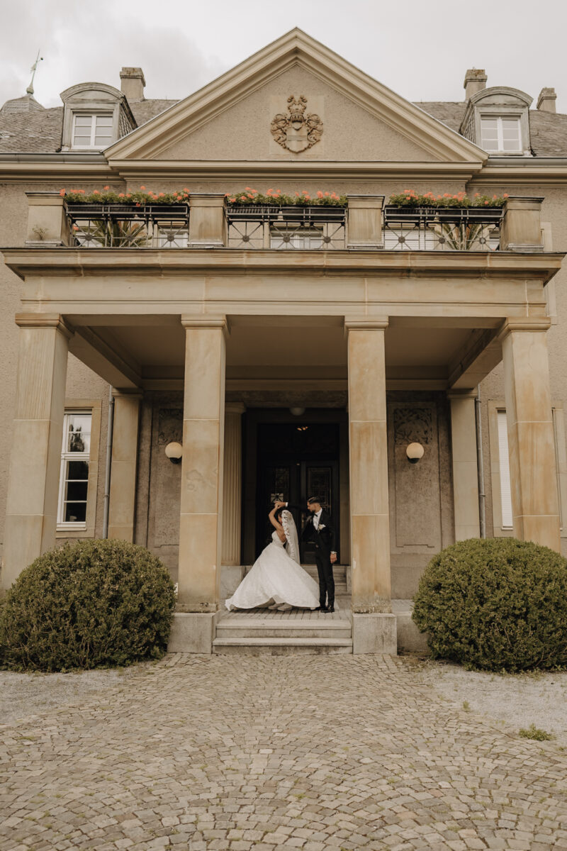 Hochzeitsportfolio, Braut wird vom Bräutigam unter seinem Arm her gedreht. Die Szene spielt sich vor enem Schlosseingang ab