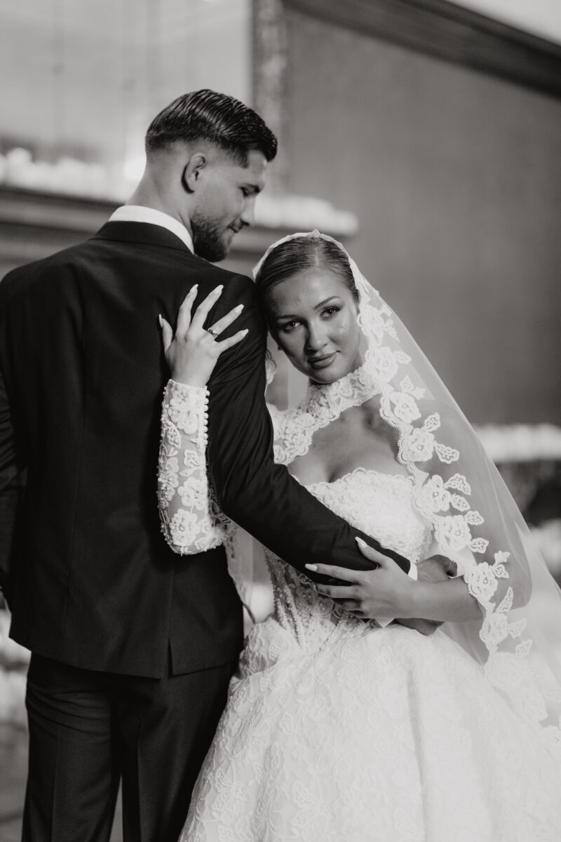 Hochzeitsportfolio, Emotionale Schwarzweißaufnahme, Braut umarmt Bräutigam von der Seite und schaut dabei in die Kamera