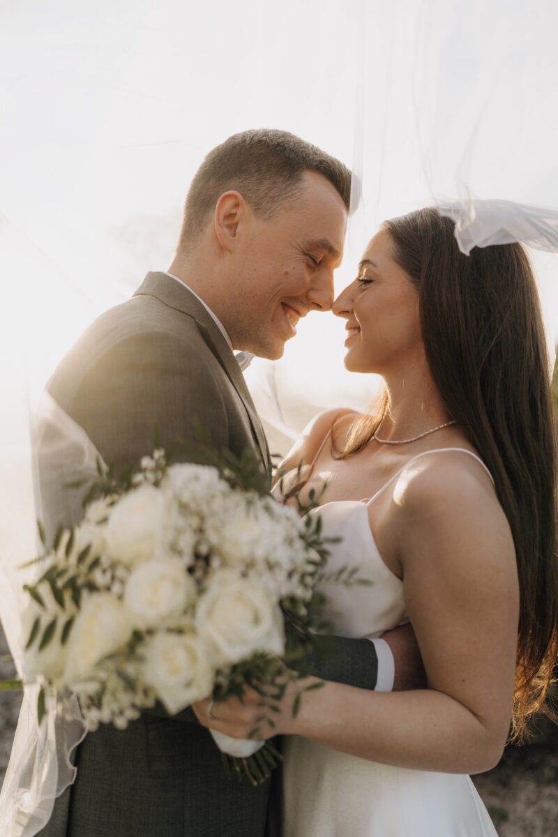 After Wedding, Braut und Bräutigam lächeln sich an, ihre Nasenspitzen berühren sich. Die Sonne scheint von hinten auf die beiden. Die Braut hält ihren Blumenstrauß