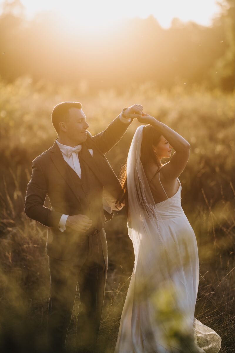 After Wedding Portfolio, Braut und Bräutigam stehen in einem Feld und tanzen zusammen. Der Bräutigam dreht seine Braut und das Sonnenlicht strahlt die beiden an.
