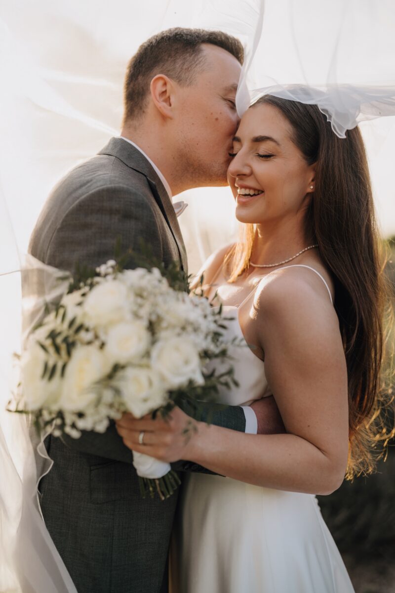 After Wedding Portfolio, Braut und Bräutigam halten sich in den Armen. Der Bräutigam küsst die Braut auf die linke Wange. Die Braut lächelt