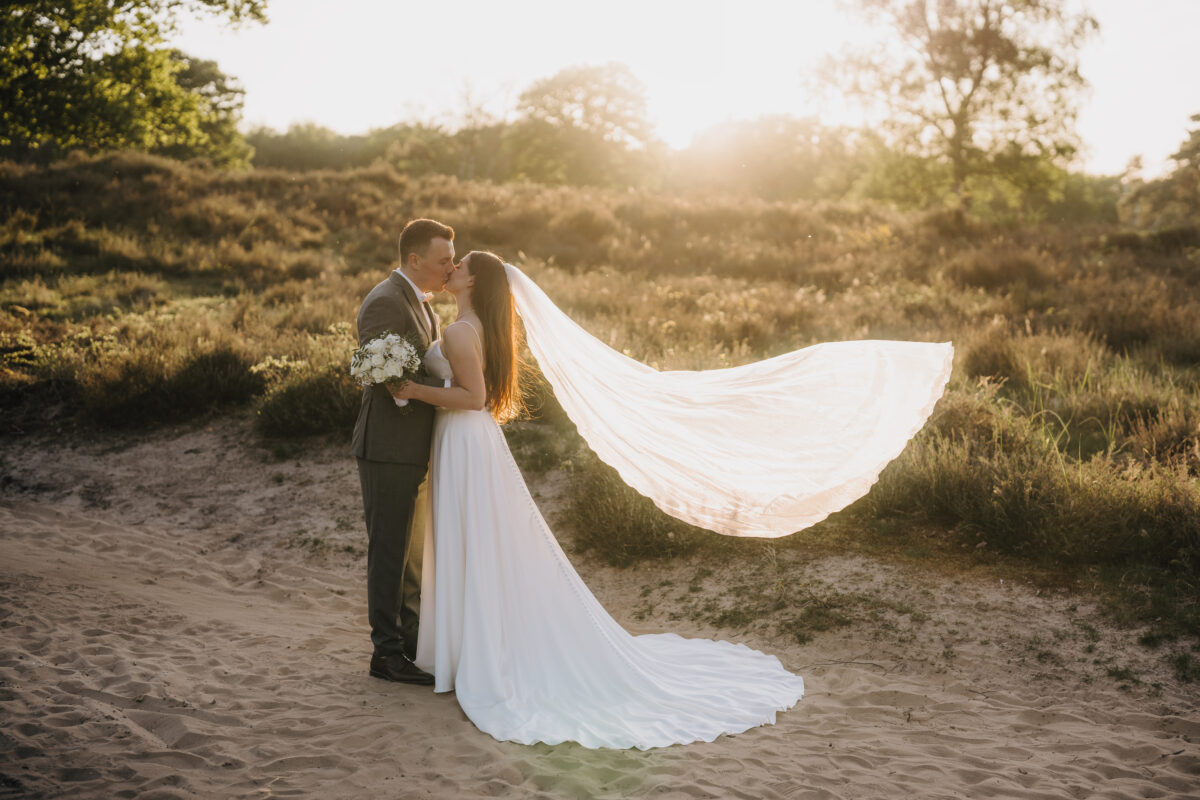 Hochzeitsportfolio, Brautpaar steht gegenüber voneinander und küsst sich, der Schleier der Braut weht im Wind, das Paar wird von hinte von der Abendsonne in ein angenehmes Licht getaucht