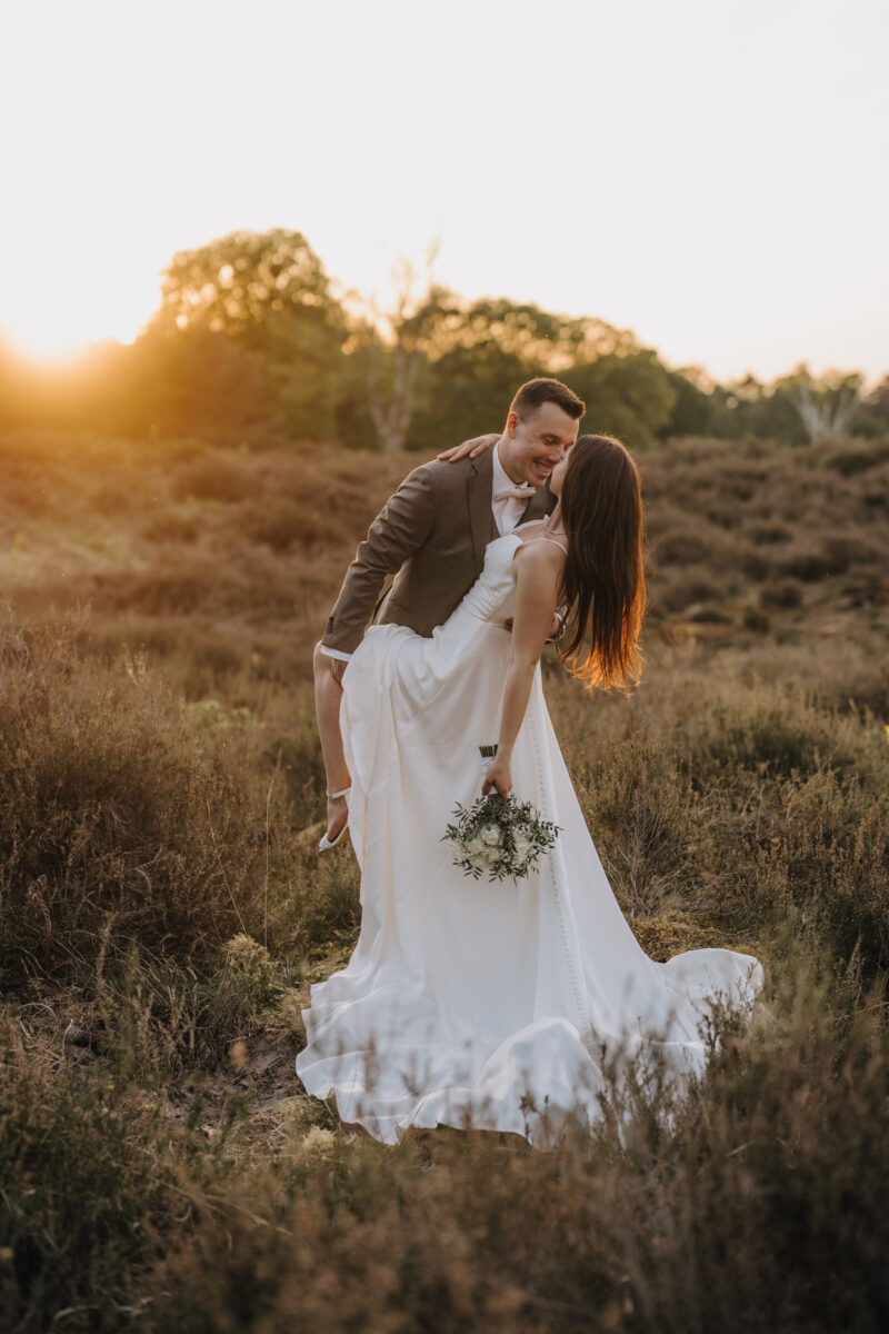 Hochzeitsportfolio, Braut und Bräutigam stehen innerhalb einer grünen Landschaft, die von der Sonne angestrahlt wird. Bräutigam lehnt sie leicht nach hinten und hält ihr Bein. Beide sind sich ganz nah und strahlen sich an.
