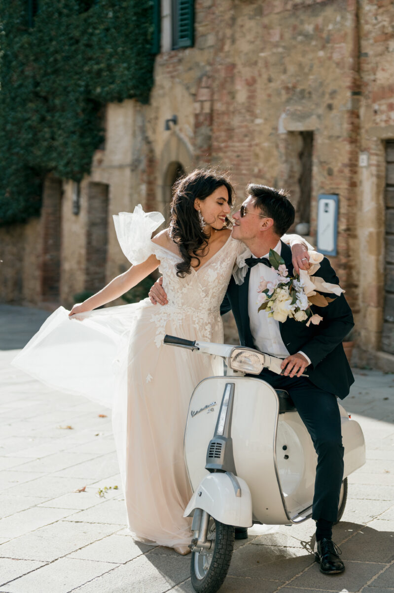 Hochzeitsportfolio, Bräutigam sitzt auf einer Vespa und hält mit der einen Hand den Lenker, mit der anderen umarmt er die Taille seiner Braut. Beide schauen sich verliebt an.