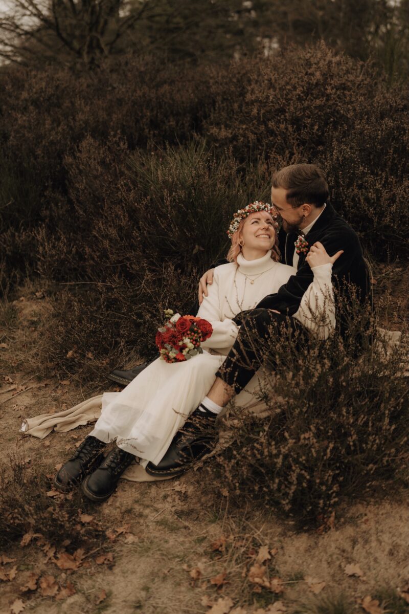 After Wedding, Braut und Bräutigam sitzen auf einer Wiese, sie liegt in seinen Armen. Beide lächeln sich an. Sie hält sich an seinem Oberarm fest.